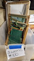 Photo of three wooden hand looms stacked inside a plastic bin, one with green and multicolored yarn woven on it.
