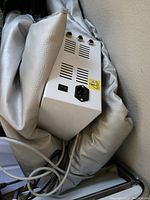 Close-up photo of the sauna blanket control unit showing power inlet, on/off switch, and three control knobs on a white rectangular unit connected to the blanket
