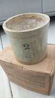 Front of large cream-colored stoneware crock with '2' and maker's stamp on the side, sitting on a cardboard box.