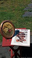 Oval framed original oil painting by Lorraine Heryet alongside MC Escher masterprint book on red stool outdoors