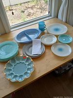Overview of all blue and turquoise dinnerware items laid out on wooden table by window. Includes divided plate, cups, plates with floral and blue dot patterns, and a turquoise deviled egg plate with gold trim.