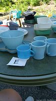 Photo of four light blue mugs and one smaller light blue mug along with a large mixing bowl in light blue glass on a table outdoors.