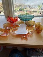 Top-down view of entire lot showing various Carnival glass bowls and dishes in iridescent orange, blue, and amber colors arranged on a wooden table by a window.