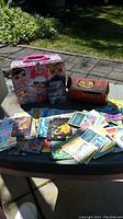 Image of metal lunch box with LOL Surprise artwork, wooden box labeled 'Anime Classic' and collectible cards spread on a table outdoors.