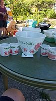 Full view of punch bowl and 4 mugs arranged on a green round table outdoors.
