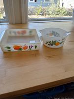 Side-by-side view of square baking dish and round mixing bowl with fruit designs on wooden surface.
