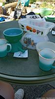 Photo showing FireKing Gay Fad white casserole dish and fruit bowl with colorful fruit motif, two ombre green mugs, and small green ribbed bowl with handle on outdoor table