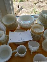 Photo shows assorted Fire-King milk glass bowls, cups and plates with gold trim stacked on wooden table in natural light.