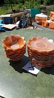 Two stacks of peach luster flower petal shaped plates and bowls on a green table outdoors. Several plates stacked on right side, several bowls stacked on left side. Background includes other orange glass items and outdoor objects.