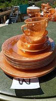 Photo showing stacked peach luster lotus W Fireking cups, saucers and plates on a green table outdoors.
