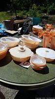 Photo showing several peach luster casserole dishes with some lids arranged on a green outdoor table.