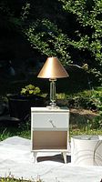 White Mid-Century Modern side table with drawer and open compartment, Lucite lamp with metallic gold-colored shade on top, set outdoors on white cloth with greenery background.
