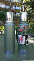 Two vintage Stanley green thermoses, one with a handle and one without, shown standing upright with metal cup lids on top. Both have worn finish and visible stickers on one thermos.