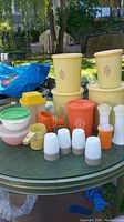 Wide view of all vintage Tupperware containers and kitchen items on outdoor glass table with grass and backyard background.