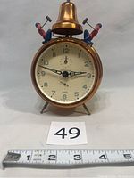 Front view of the brass alarm clock showing clock face, bell, and figurines.