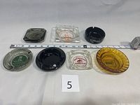 All seven ashtrays shown laying flat with measuring tape for scale and number 5 plate, showing different colors and logos.