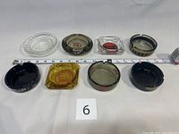 Photo showing all eight vintage hotel and casino ashtrays laid out in two rows on a white surface with a measuring tape, labeled with a card numbered 6.