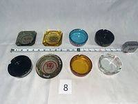 Eight vintage glass casino ashtrays arranged with measuring tape for scale. Varied colors include smoke gray, amber, black, blue, and clear glass. Ashtrays have printed casino logos including Riverboat Casino, Bonanza Casino, Harveys, and others.