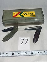 Two folding pocket tools, one with green handle and one with wood handle, displayed with plastic box for sharpening stone, measuring tape shown for scale
