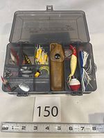 Open small transparent plastic fishing tackle box showing various vintage fishing lures and accessories inside, including Rapala lures, metal spinners, hooks, spoons, wooden bobber, and leather sheath pouch. Labeled with lot number and ruler for size reference.