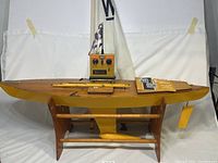 Side profile of the China Boy remote sailboat on wooden stand showing the wooden deck, yellow hull, mast, sail with 'W' marking, and Kraft radio controller on top with manual.