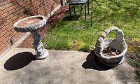 Wide shot showing both the cement birdbath and the basket planter on a concrete patio near grass and a brick wall.