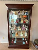 Front view of the tall, dark wood collectors cabinet showing four glass shelves filled with porcelain figurines. Mirrored back inside the cabinet.