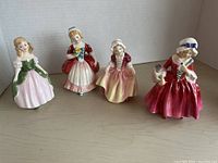 Four Royal Doulton porcelain figurines showing front views on shelf, wearing period dresses in pastel and red tones.
