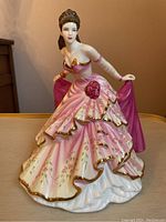 Front view of Grace figurine showing detailed facial features and layered pink gown with gold trim and floral accents, holding a pink shawl.