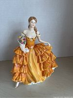 Front angle of the figurine showing the orange and yellow layered dress and the bouquet with card in hand.