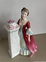 Front angled view of the Royal Doulton figurine showing the woman in a red and pink dress next to a white pedestal with flowers.
