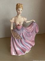 Front view of Royal Doulton Rachel figurine showing detailed face, off-shoulder pink/lilac gown, fan in left hand and dress lifted with right hand.