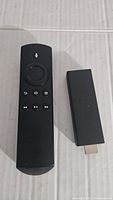 Amazon Fire TV Stick streaming device alongside its remote control on a cardboard surface, showing full device and control buttons.