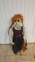 Full body view of the creepy Halloween girl prop standing barefoot on tile floor with long black dress, pale green cracked face, and holding a brown teddy bear.