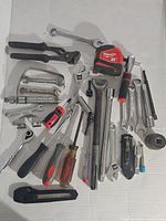 Full view of assorted hand tools scattered on white background including Milwaukee measuring tape, pliers, wrenches, screwdrivers, ratchet handles, sockets, utility knife, small hacksaw.