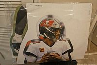 Cardboard cutout showing Tom Brady in full Buccaneers uniform with helmet, holding a football as if preparing to throw.