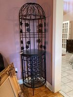Front view of metal bar rack showing intricate metal design with flowers and leaves on tall cage-like structure topped with dome, standing next to wall.