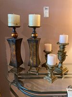 Set of three ornate resin candle holders with detailed leafy carvings and square bases, each holding a white pillar candle, along with two tall flared tin candle holders with a metallic bronze-gold finish and round candle plates, placed on glass surface.