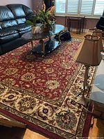 Overall living room view showing large rectangular wool carpet in deep red with ornate floral beige border under glass top coffee table, beside black leather sofa and brass lamp.