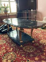 Side angle view of two tier coffee table showing black resin top and lower shelf with gold metal legs and decorative leaf details. Table on patterned carpet.