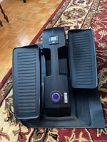 Full view of the Cubii under desk elliptical machine on the floor mat with foot pedals, display screen, and resistance knob visible.