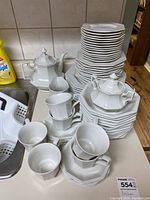 Overall view of the dish set, showing stacks of plates, cups, two teapots and bowls arranged near a kitchen sink.