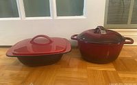 Two red cooking vessels: a round KitchenAid enamel cast iron dutch oven with lid and a rectangular Corningware ceramic Hearthstone dish with lid.