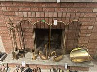 Brass fireplace screen in front of a brick fireplace with four-piece brass tool set on stand, brass log holder, two brass swans, and a brass match holder on the hearth.