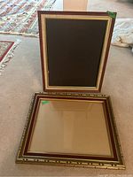 Two wood picture frames displayed on carpet; one upright frame with brown fabric matting, one horizontal frame with gold ornamental accents, red velvet matting, and glass.