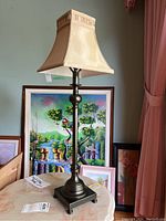 Full view of the candlestick style lamp with square fabric shade on a round table, artwork in the background.