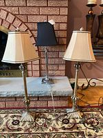Three table lamps shown together: two matching gold-tone metal base lamps with beige fabric shades and one smaller silver-tone metal base lamp with black shade.