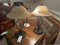 Pair of metal and glass table lamps with frosted beige bell-shaped shades and dark metal bases on a wooden table, along with the metal goose neck desk lamp in background.