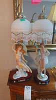 Pair of two lamps with white frosted glass shades featuring beaded fringe, each on wooden base with porcelain female figurines.