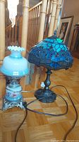 Photo showing both lamps side by side on wooden floor near staircase. Blue stained glass lamp on right, white glass lamp on left.